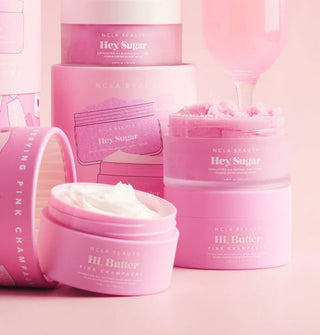 Opened pink jars of NCLA Beauty Hi, Butter moisturizer and Hey, Sugar scrub with other unopened jars, gift tubes, and a glass of pink champagne in the background