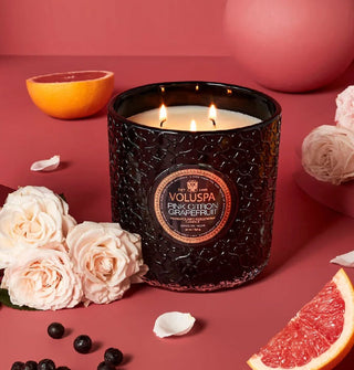Lit three-wick Pink Citron Grapefruit Voluspa candle in a black glass vessel is staged on a pink backdrop with roses, berries, and slices of grapefruit