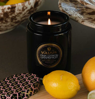 Lit Pink Citron Grapefruit Voluspa candle in black glass jar is staged with its corresponding lid, citrus fruits, and speckled brown dishes