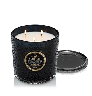 Lit three-wick Pink Citron Grapefruit Voluspa candle in a black glass vessel with matching lid which has been removed and set to one side