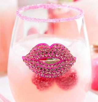 A clear stemless wine glass featuring a pink lips decal fully bejeweled in pink rhinestones is filled with a milky beverage garnished with raspberries and a pink sugared rim