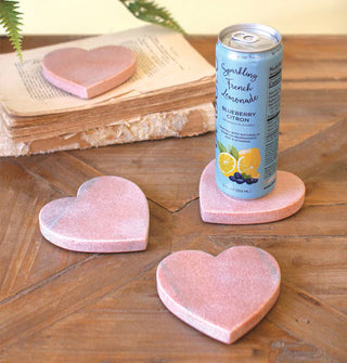 Grouping of three pink heart-shaped marble coasters on a wooden tabletop with one slim can placed on one and a fourth coaster placed atop a stack of two distressed books near fern branches