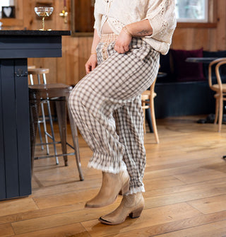 Model kicking up leg wears a pair of plaid bloomers in a neutral palette with lace trim paired with a pair of cowgirl boots