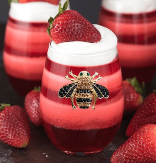 Clear stemless wine glass featuring a bumblebee decal bejeweled with rhinestones is filled with a red layered beverage topped with whipped cream and a strawberry