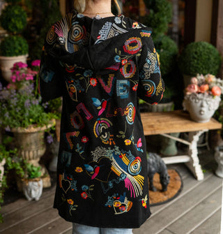 Model standing against a backdrop of potted plants and a bench on a porch deck  with back to camera wears a long black hooded cardigan with colorful embroidered designs all over it
