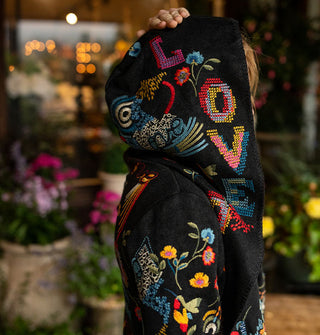 Model standing against a backdrop of potted plants against a window wears a black hooded cardigan with colorful embroidered designs all over it