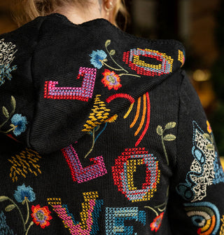 Model with back to camera wears a black hooded cardigan embroidered with colorful designs and the word, "Love" in large lettering
