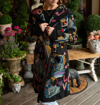 Model standing against a backdrop of potted plants and a bench on a porch deck wears a long black cardigan with colorful embroidered designs all over it