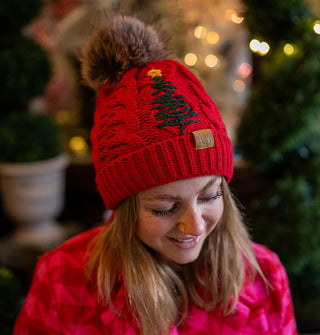 Smiling model wears a red cable-knit beanie with green tree embroidery and faux fur pompom on top