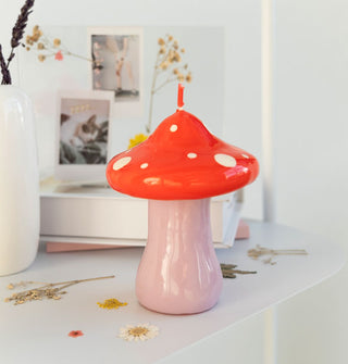 Mushroom candle with purple base and red cap with white spots sits on a ledge scattered with dried flowers