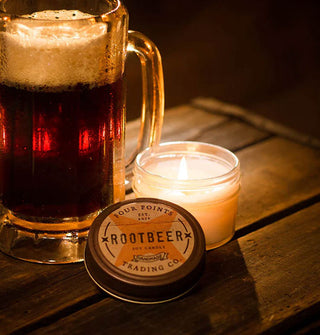 A lit Root Beer scented jar candle with lid removed is staged on a wooden surface next to a glass mug of frothy, dark soda