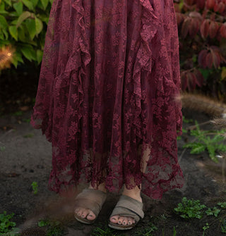 Model standing among lush greenery wears a midi-length wine red lace handkerchief skirt with ruffled edges