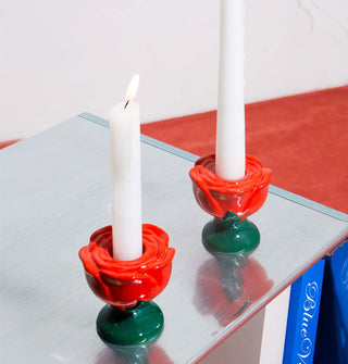 Two ceramic red rose candle holders each hold a white taper on a reflective surface