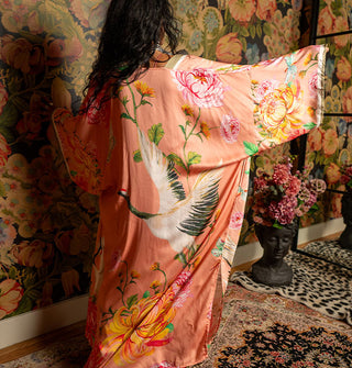 Model with back to camera and arms outstretched wears a coral-pink full-length robe with wide sleeves and a bold floral print around a large crane with wings spread