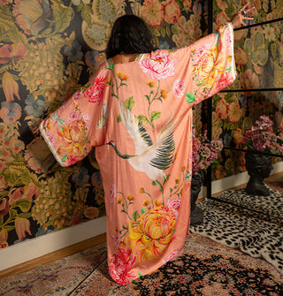 Model with back to camera and arms outstretched wears a coral-pink full-length robe with wide sleeves and a bold floral print around a large crane with wings spread