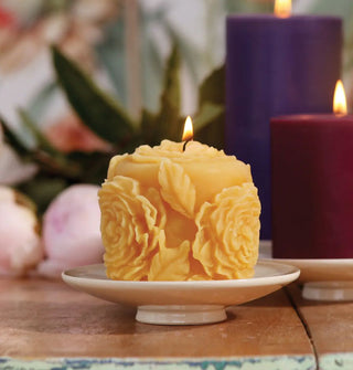 Lit sculptural beeswax rose candle on a small white pedestal dish with other lit pillar candles in the background