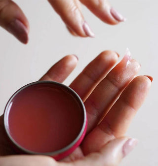 Model with a glob of salve on the tip of middle finger holds an opened round tin of the product in the palm of the same hand