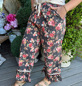 Model wears a pair of cropped black capri-style, ruffle-trimmed pants with floral print, side pockets, and white waist tie
