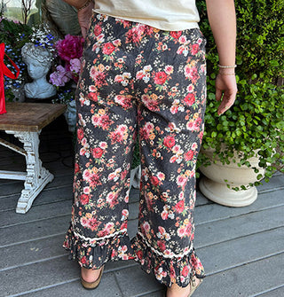 Model wears a pair of cropped black capri-style, ruffle-trimmed pants with floral print and contrastic white ric rac border