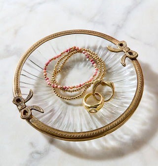 Round ribbed glass dish with brass rim featuring bow accents on either side rests on a marble tabletop holding bracelets and rings