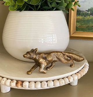 Brass running fox figurine on a wooden bead-border pedestal with a potted plant and framed artwork in the background