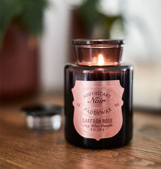 A lit Apothecary Noir Paddywax Saffron Rose candle shines from inside a black apothecary-style jar on a wooden surface with houseplants in the background