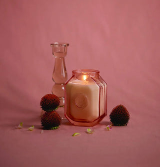Lit candle in octagonal pink glass jar is staged with botanical elements and a matching glass taper holder behind against a pink backdrop