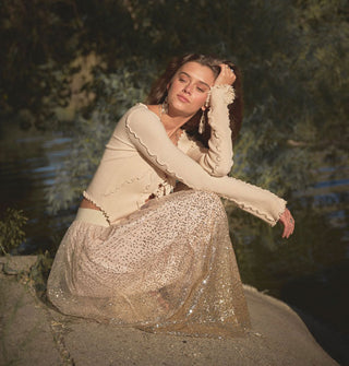 Model sitting on a rock wears a champagne-colored full-length sequined skirt against a backdrop of water and trees