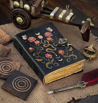 Journal with rough page edges and a black cover featuring illustration of a snake, flowers, moths, and centipedes rests on a burlap-covered wooden surface with antique wood and brass implements