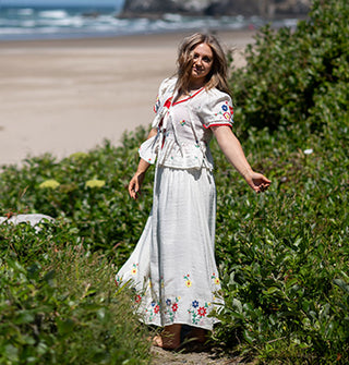 Model standing along the coast wears a white top and skirt featuring colorful floral embroidery