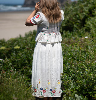 Model standing along the coast wears a white skirt and peplum top, both featuring colorful floral embroidery