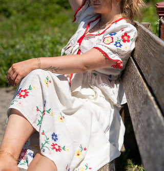 Model reclining on a wooden bench wears a matching white skirt and top set, both with colorful embroidery and trim