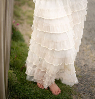 Model standing in bare feet beside a wooden fence wears a cream-colored tiered ruffle maxi skirt
