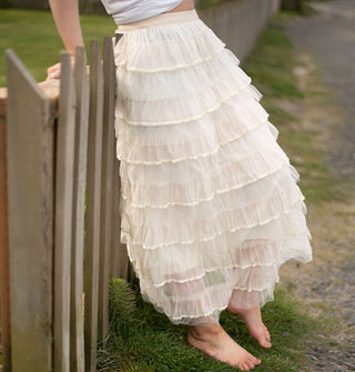 Model leaning against a wooden fence wears a cream-colored tiered ruffle maxi skirt