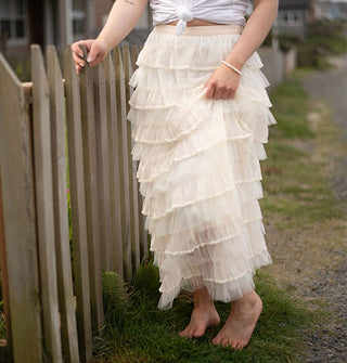Model standing in bare feet beside a wooden fence wears a cream-colored tiered ruffle maxi skirt