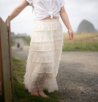 Model standing in bare feet beside a wooden fence wears a cream-colored tiered ruffle maxi skirt against a seaside backdrop
