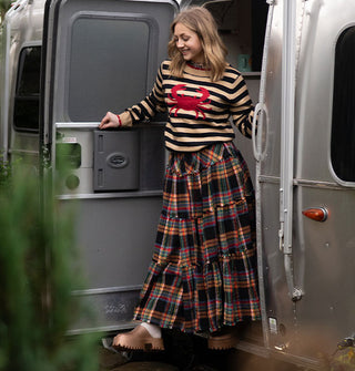 Smiling model stepping out of an Airstream camper wears a beige and black striped sweater with red crap emblem on the front paired with a multicolored plaid maxi skirt and copper clogs