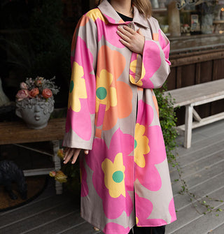 Model wears an overcoat with large floral print in a gray, pink, orange, yellow and green color palette