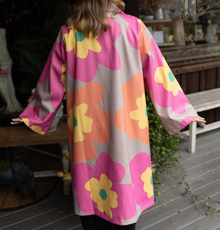 Model wears an overcoat with large floral print in a gray, pink, orange, yellow and green color palette