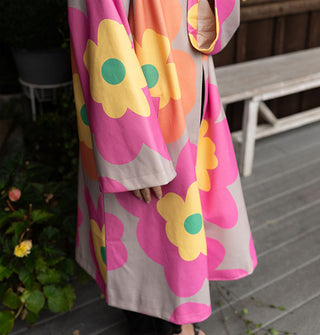 Model wears an overcoat with large floral print in a gray, pink, orange, yellow and green color palette