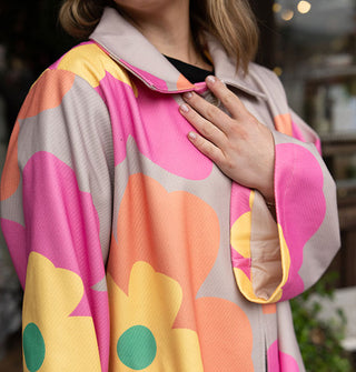 Model wears an overcoat with collar and large floral print in a gray, pink, orange, yellow and green color palette