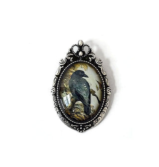 Oval brooch features illustration of a crow under clear glass in an ornate antiqued silver fame