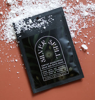 Black packet of Silver Spell Earthy Eucalyptus Dead Sea Bath Soak with dark green floral and fern motif and white lettering rests on a terracotta-colored surface scattered with coarse salt