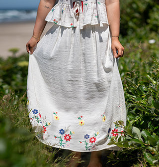 Model standing along the coast wears a white top and skirt featuring colorful floral embroidery
