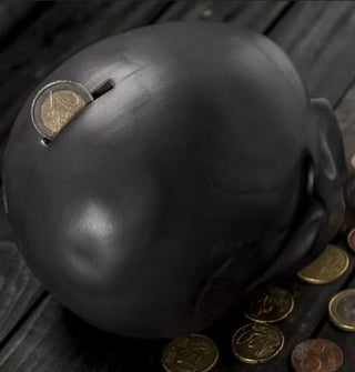 Black skull-shaped money bank is staged with a coin emerging from its top slot with other coins strewn at its base on a black wooden tabletop