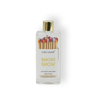Clear rectangular glass bottle with gold cap and a label that says, “Smoke show” in gold lettering contains multicolor-tipped wooden matchsticks