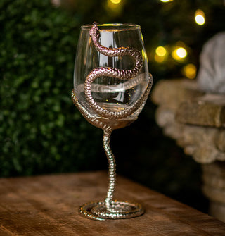 Wine glass with a clear bowl and silver snake wrapped partially around it, forming the stem and base with its body rests on a wooden tabletop in front of a light-strung garden backdrop