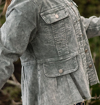 Model wears a stone wash-effect blue-gray corduroy peplum jacket against a grassy backdrop