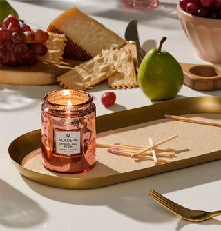 Lit speckled pink glass Voluspa Sparkling Rose candle rests on a matte gold tray with wooden matchsticks in front of charcuterie in the background and a gold fork in front
