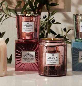 Speckled pink glass Voluspa Sparkling Rose candle jars and matching metallic box in front of an olive branch